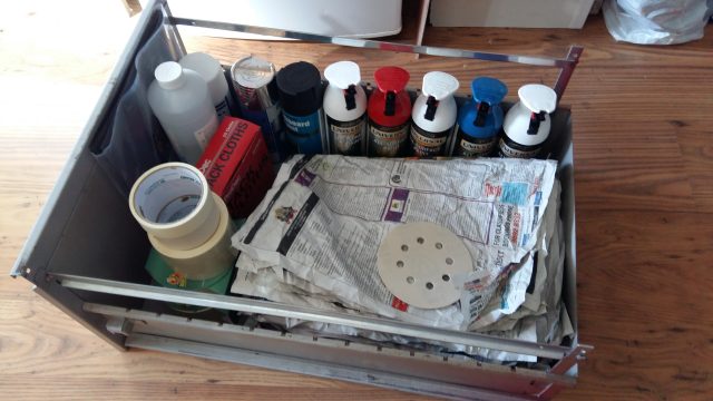 An image of a filing cabinet drawer, placed on a wooden laminat floor, and filled with spray paint and other craft materials.