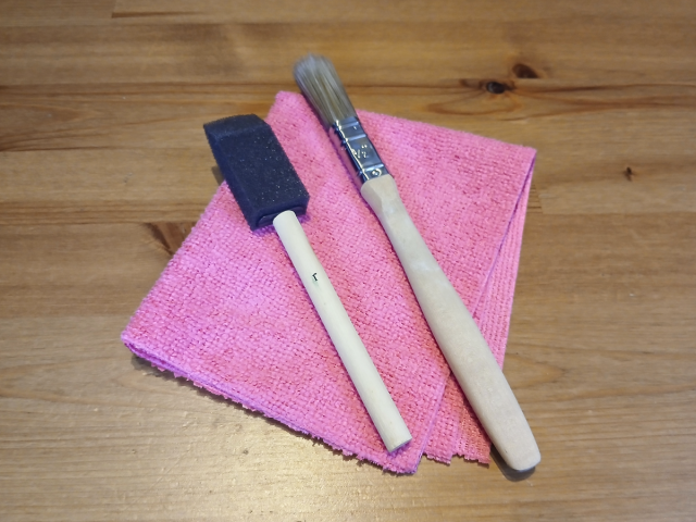 Image shows a one inch foam brush, next to a half inch bristle brush, placed on a pink microfibre cloth. Shown with a wooden background.