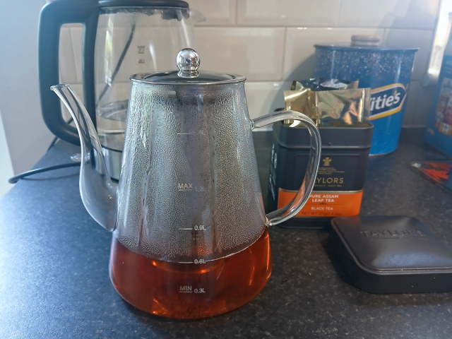 Image shows a glass teapot on a kitchen work surface, with an open tea caddy to the right-hand side.