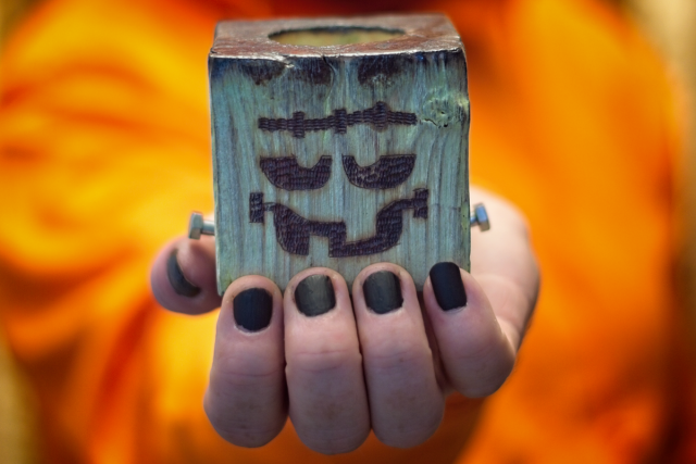 Image shows a hand holding a wooden tealight holder, stained white, with the face of Frankenstein's Monster burnt into the front. Shown on an orange background.