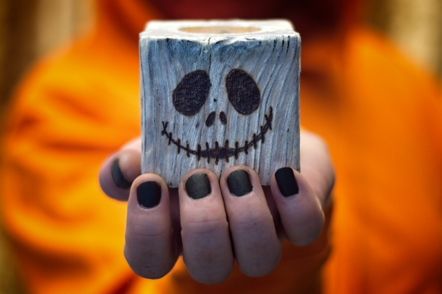 Image shows a hand holding a wooden tealight holder, stained white, with the face of Jack Skellington burnt into the front. Shown on an orange background.