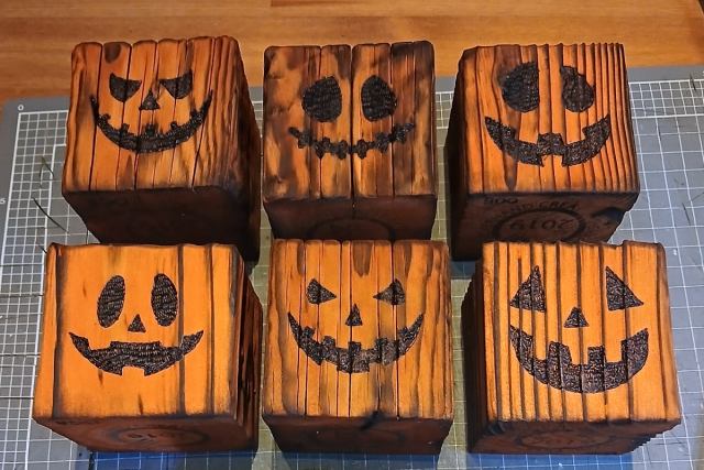 Image shows six handcrafted, wooden pumpkin tealight holders, with pumpkin faces burnt into the surface. Shown on a grey and wooden background