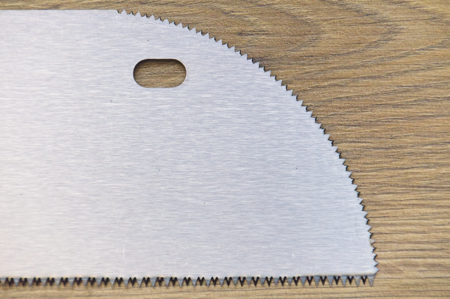 A close-up of the the saw teeth on the rounded nose of a floorboard saw. Shown on a wooden background.